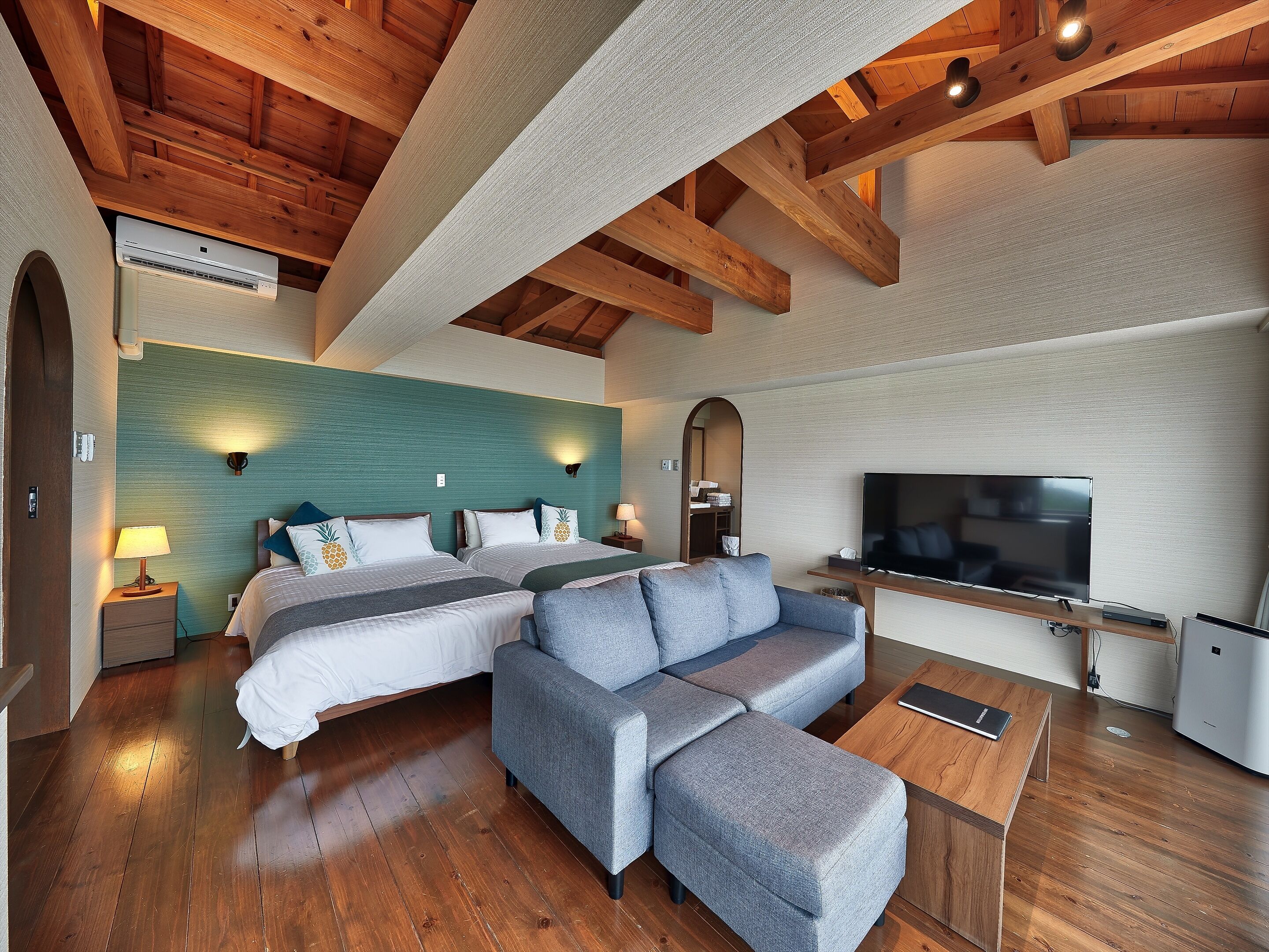 Habitación de hotel moderna con dos camas, un sofá gris, televisor de pantalla plana, piso de madera y estructura de techo de madera.