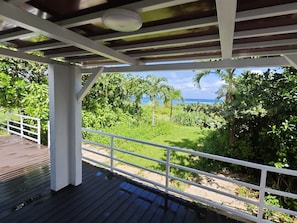 A covered outdoor area with a wooden deck and white railings, overlooking a lush garden and a view of the ocean. - Nagahama Beach Resort Kanon (Nakijin)