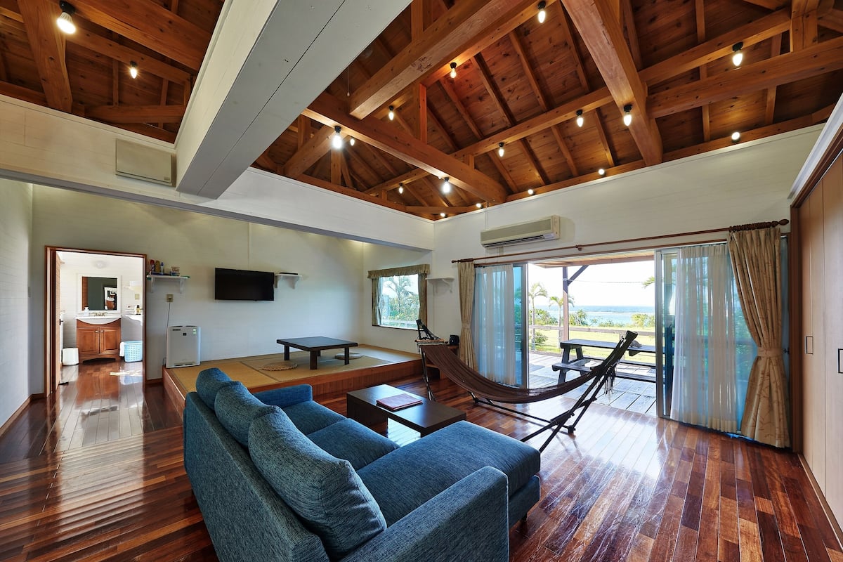 A spacious living room with a wooden ceiling, a blue sofa, a table, and a hammock.
