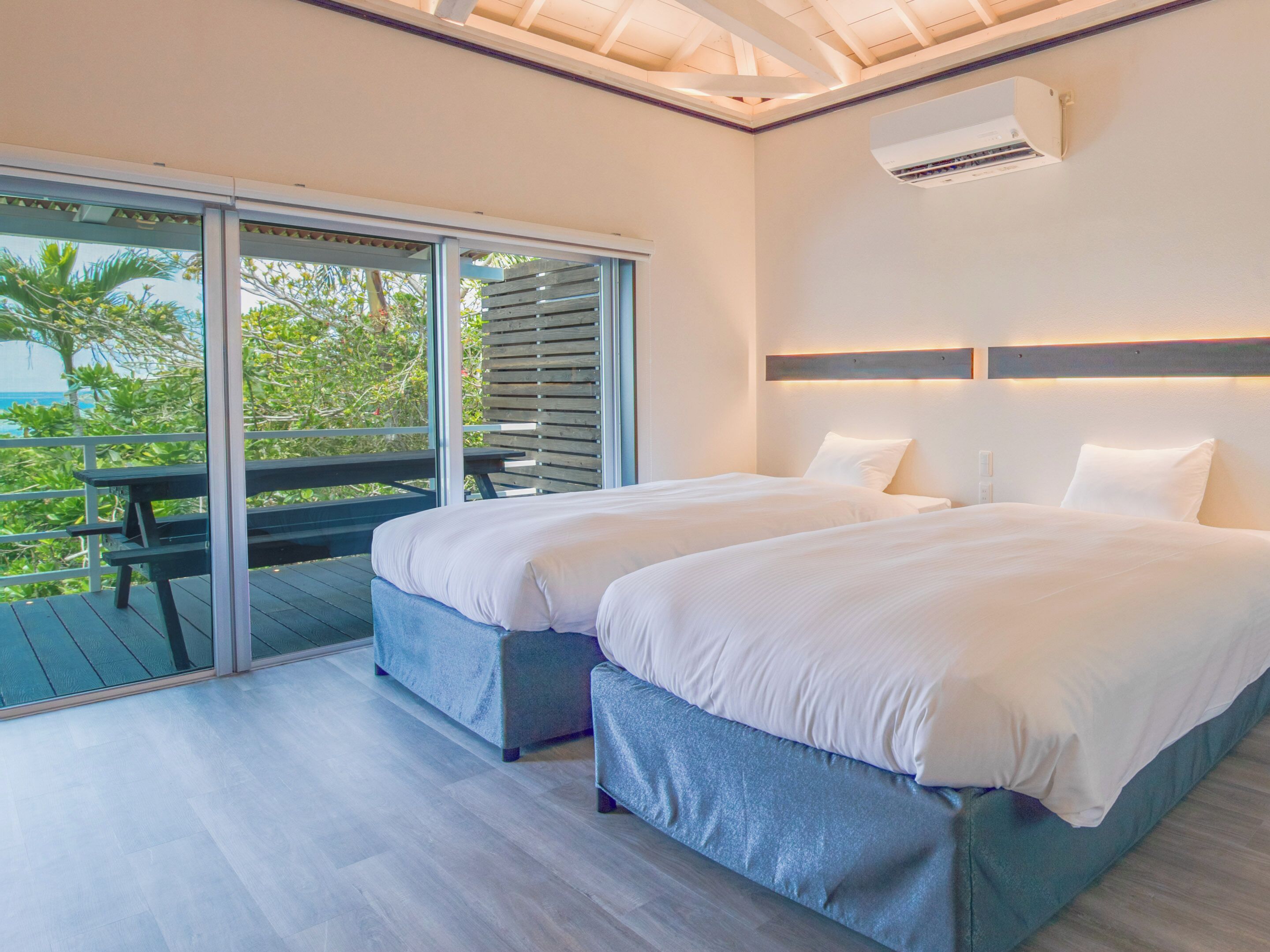 A modern hotel room with two beds, a balcony with a table, and a view of greenery.