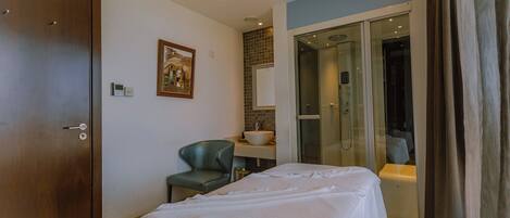 A hotel room with a bed, a chair, a bathroom with a glass door, and a painting on the wall.