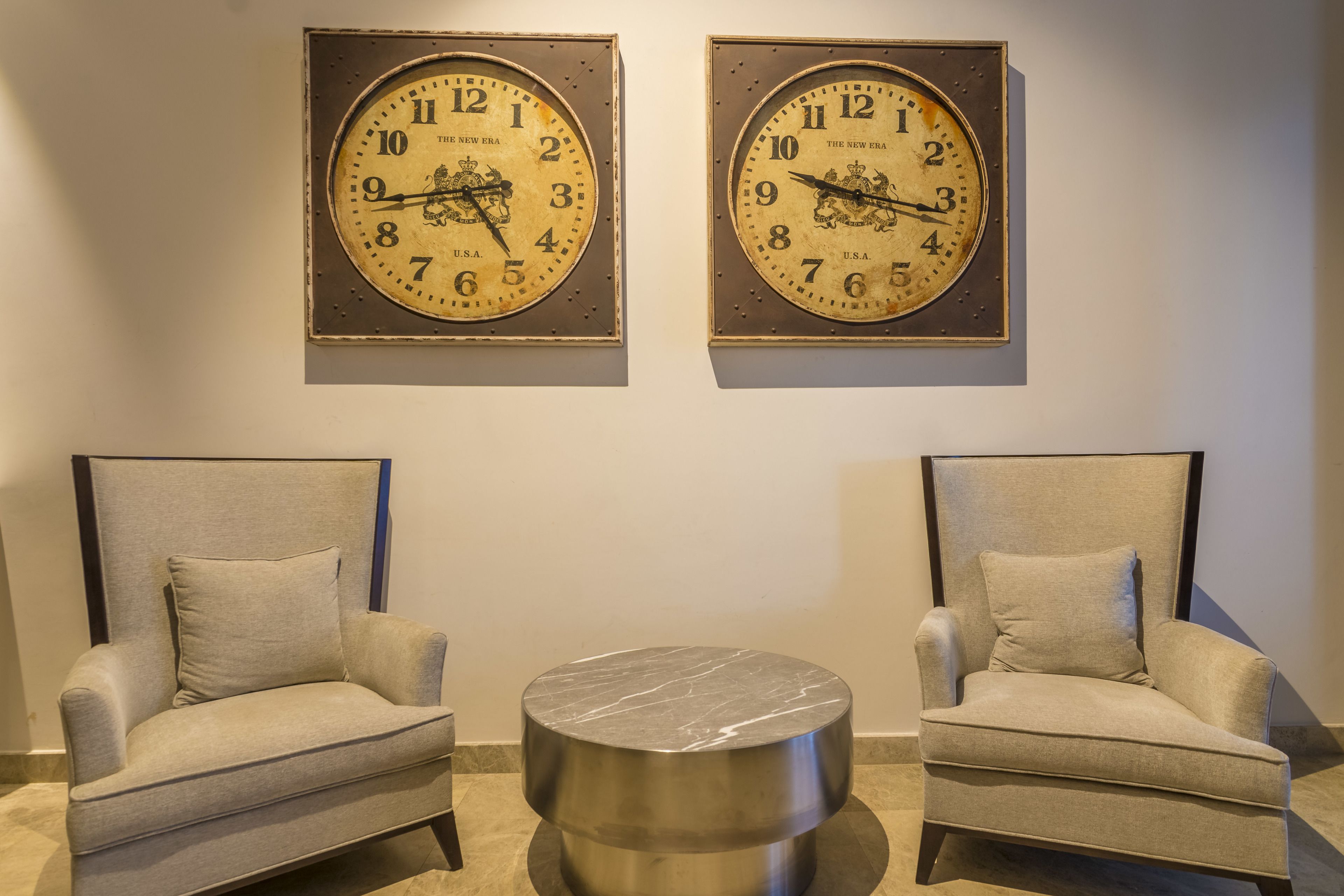 Two armchairs and a round table in a room with two clocks on the wall.