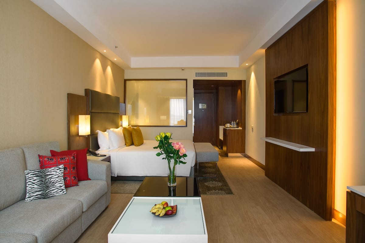 A modern hotel room with a sofa, a bed, a coffee table with fruit, a TV, and a mirror.