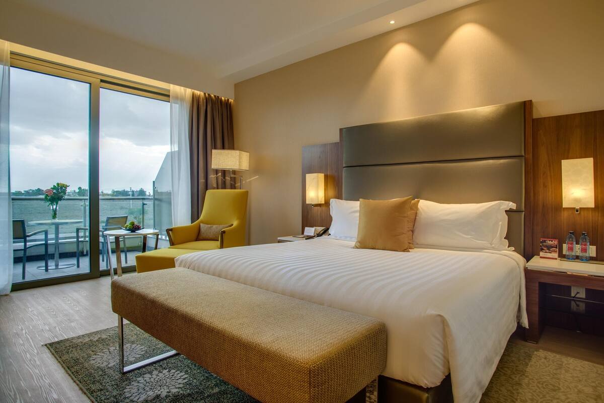 A hotel room with a large bed, a bench, a desk with a lamp, a chair, and a view of the outdoors.