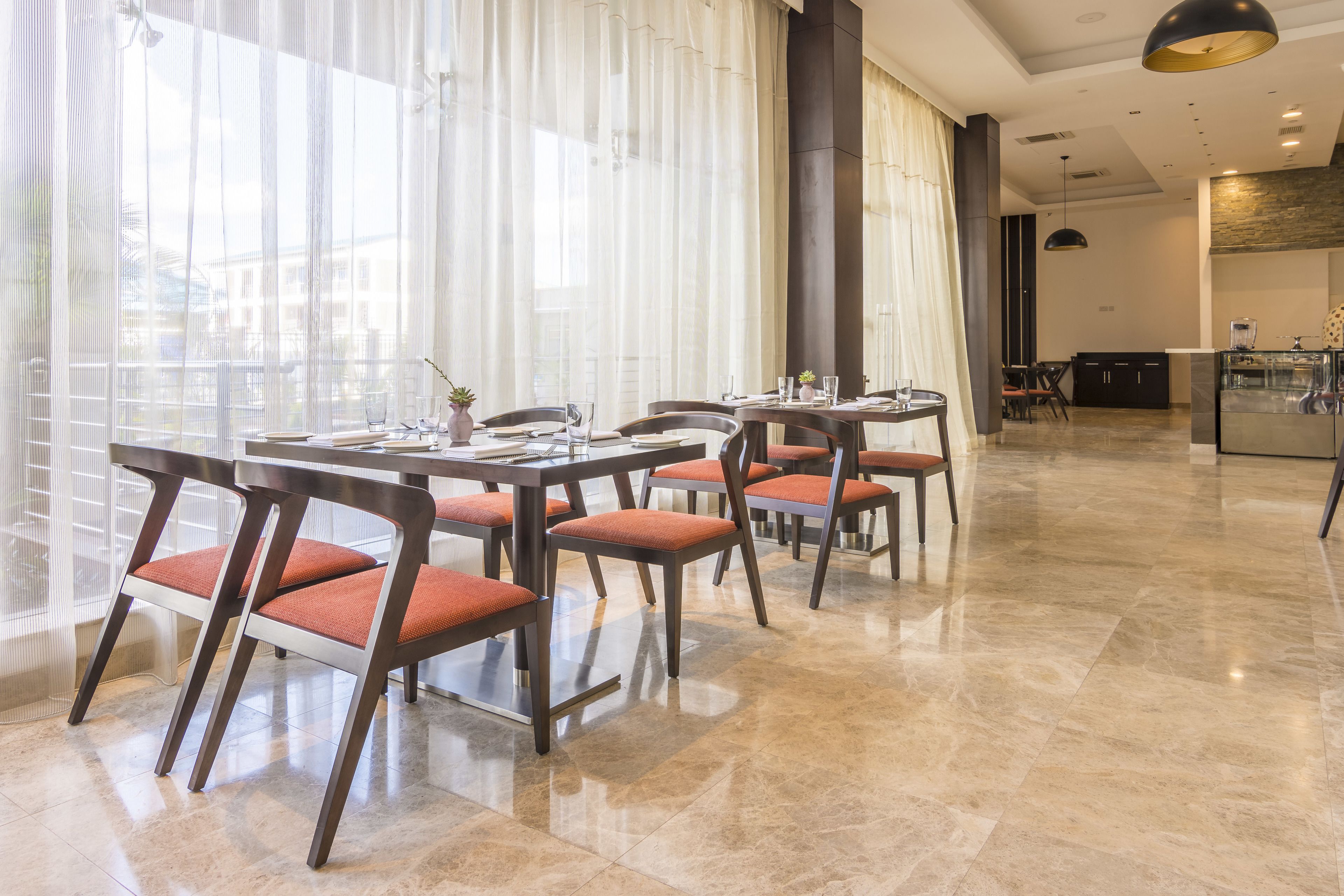 Un comedor amplio con varias mesas y sillas, ventanas grandes con cortinas translúcidas y una cocina moderna al fondo.