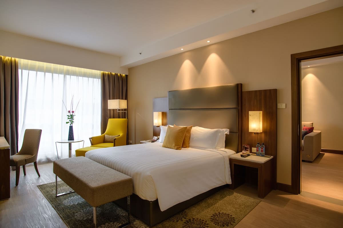 A hotel room with a large bed, a bedside table with lamps, a chair, and a small table with a vase.