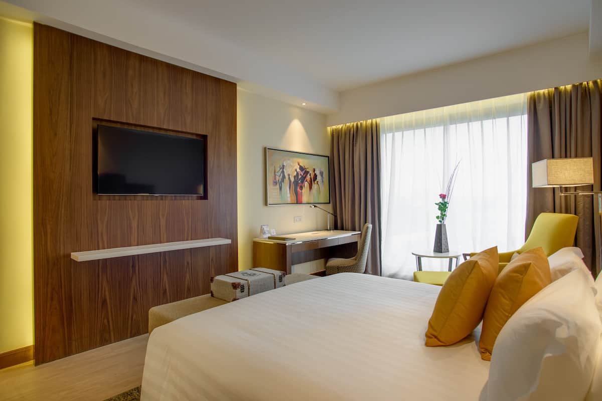 A hotel room with a large bed, a desk with a chair, a flat-screen TV mounted on the wall, a painting on the wall, and a window with curtains.