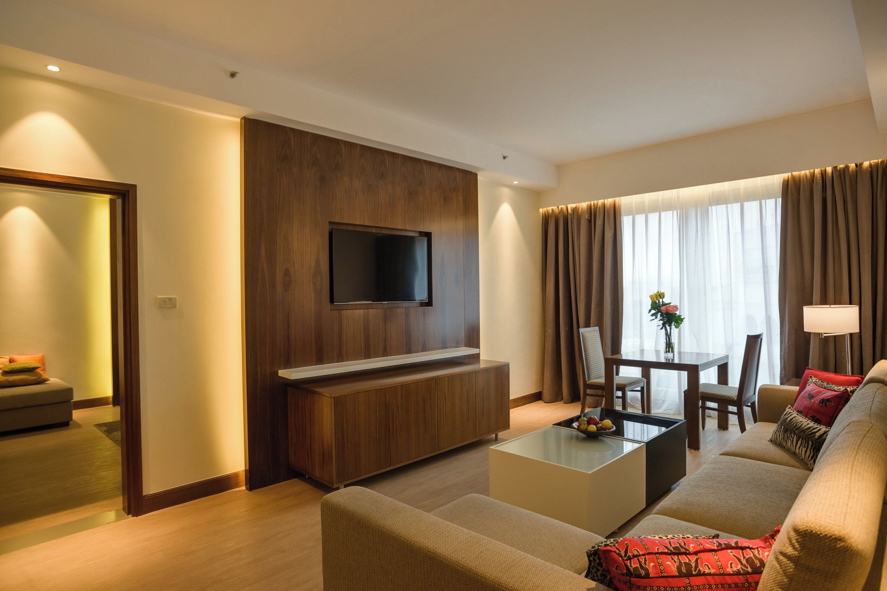 Een moderne hotelkamer met een flatscreen televisie bevestigd aan een houten paneel, een bank, een salontafel, en een zithoek met een tafel en stoelen.