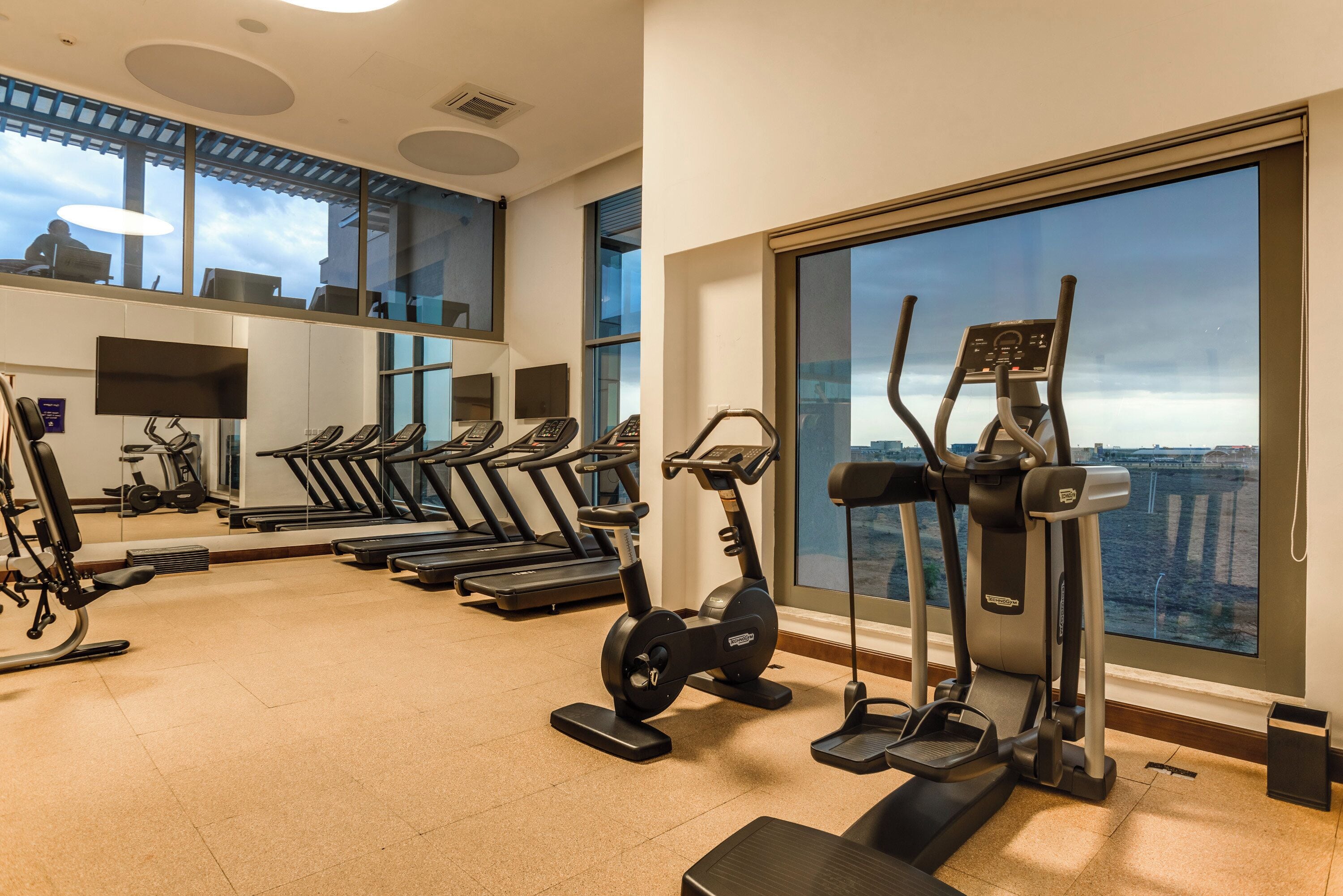 A gym with treadmills, elliptical machines, and a stationary bike, with large windows offering a city view.