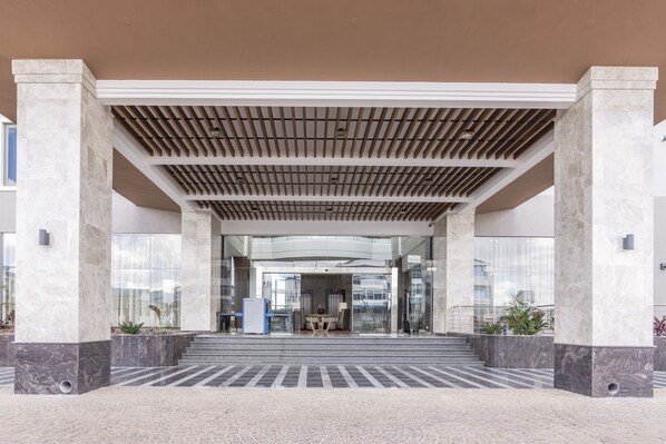 Una entrada de edificio moderno con una galería techada, columnas de mármol y un área de descanso con una mesa y sillas.