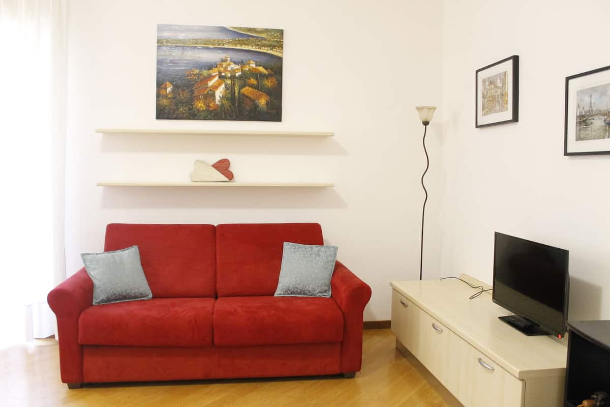 A red sofa with two pillows, a TV on a stand, and artwork on the wall.