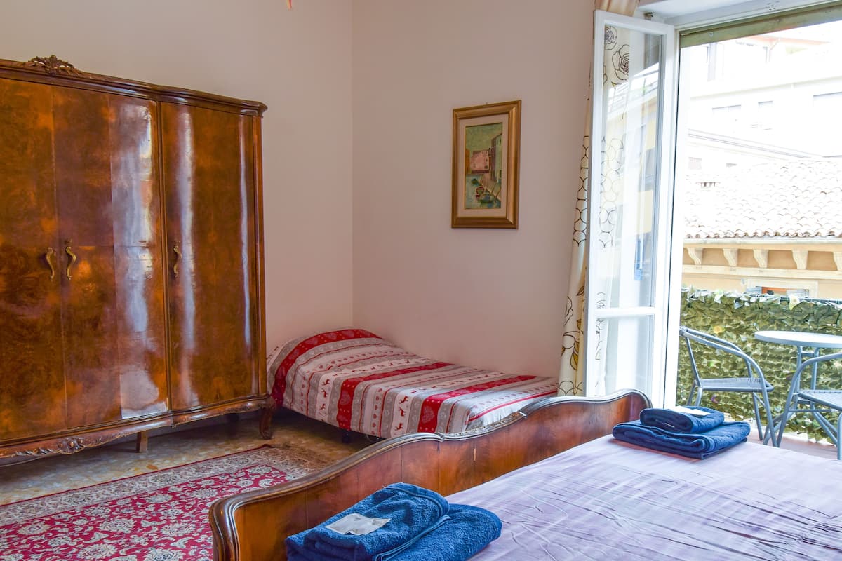 A bedroom with a wooden wardrobe, a bed with a red and white patterned bedspread, a blue towel on the bed, and a window with a view of buildings outside.