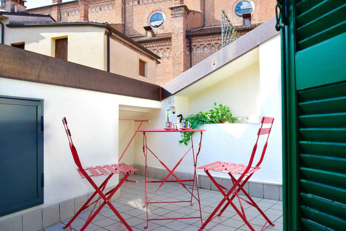 A small outdoor balcony with red metal chairs and a table, a potted plant, and a view of a brick building in the background.