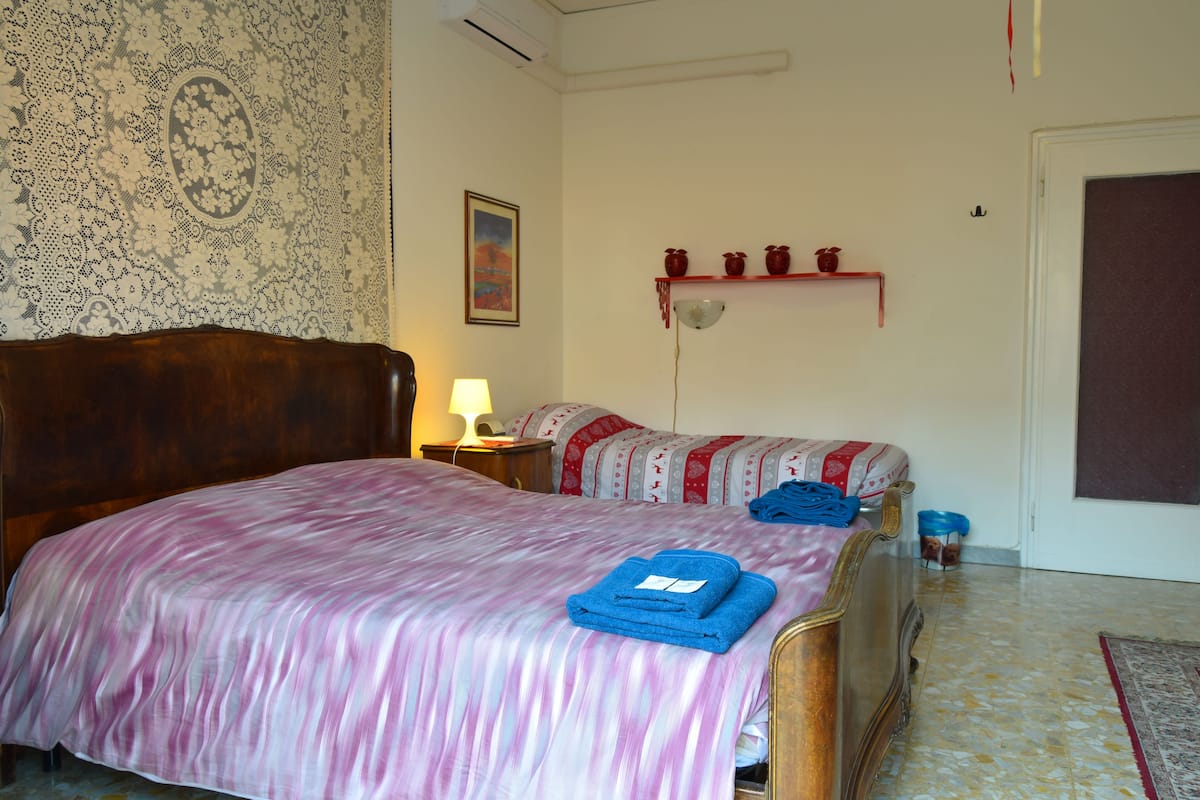 A bedroom with a large bed, a bedside table with a lamp, a wall tapestry, a shelf with decorative items, and a door.