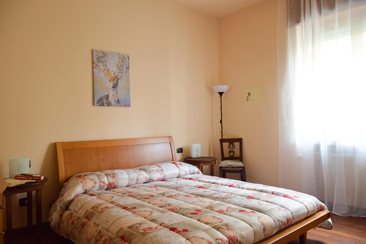A bedroom with a wooden bed, a bedside table with books, a lamp, and a painting on the wall.