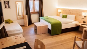 Ein Hotelzimmer mit einem großen Bett, einem kleineren Bett, einem Schreibtisch mit Stuhl, einer hölzernen Bank, einem Fernseher und einem Spiegel.
