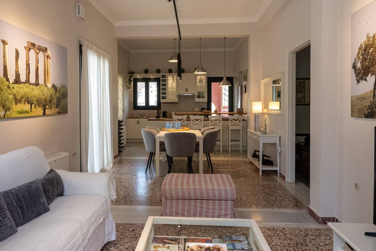 A spacious living area with a sofa, a coffee table with magazines, a dining table with chairs, and a kitchen in the background.