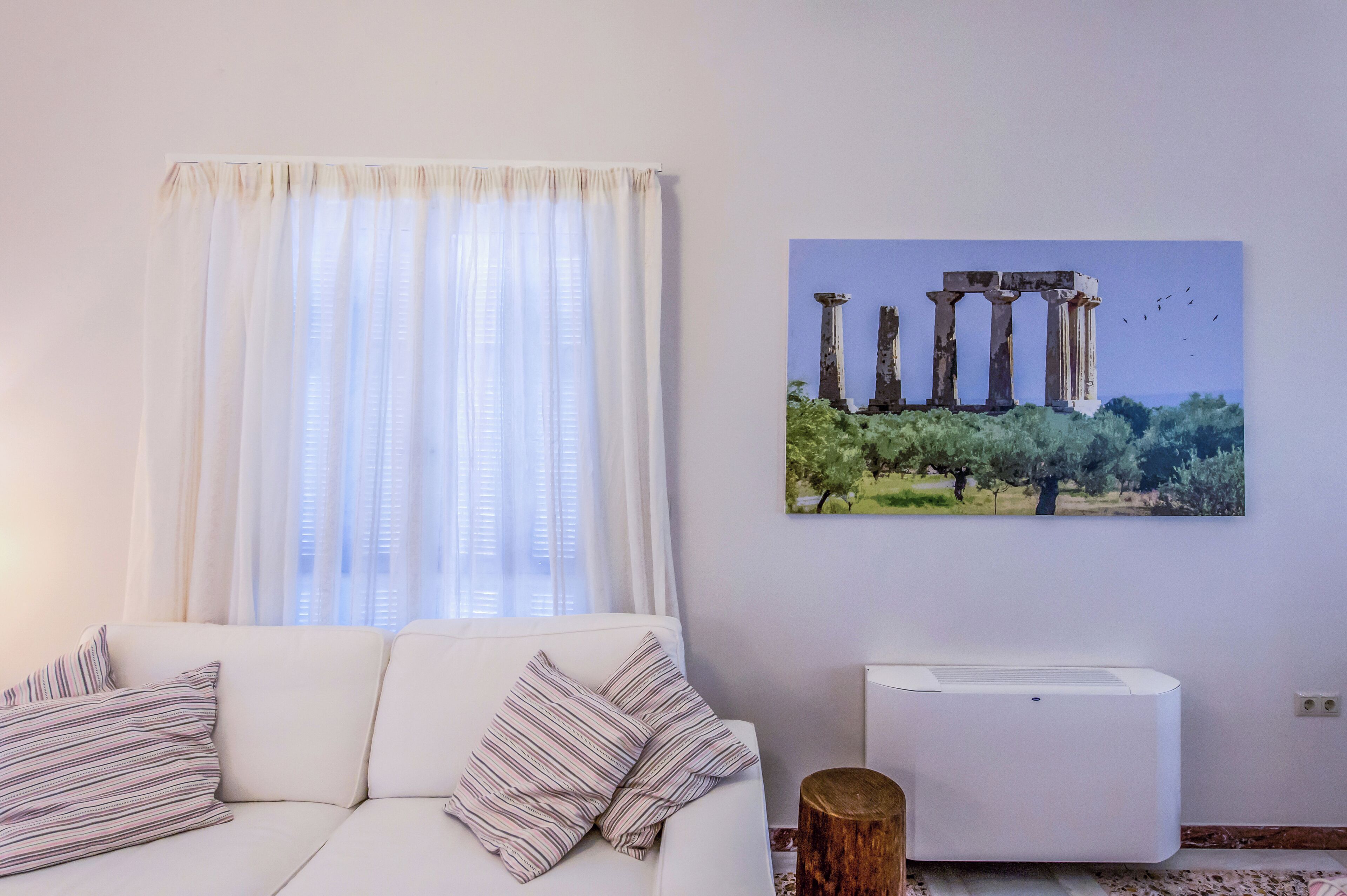 Een woonkamer met een witte bank, gestreepte kussens, een raam met dunne gordijnen en een luchtverwarmer aan de muur.