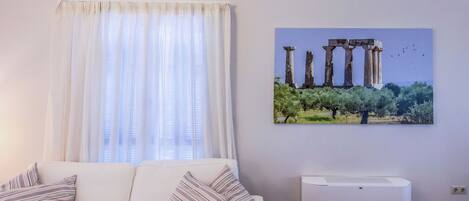 Een woonkamer met een witte bank, gestreepte kussens, een raam met dunne gordijnen en een luchtverwarmer aan de muur.