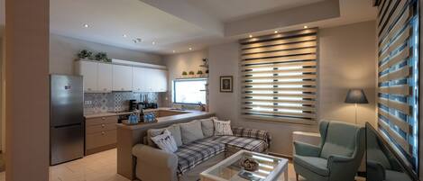 Una sala de estar moderna con sofá, mesa de centro y televisor. Hay una zona de cocina con armarios y un refrigerador.