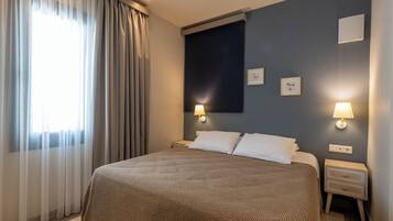 A hotel room with a large bed, two bedside tables with lamps, a dark wall with framed pictures, and sheer curtains on the window.