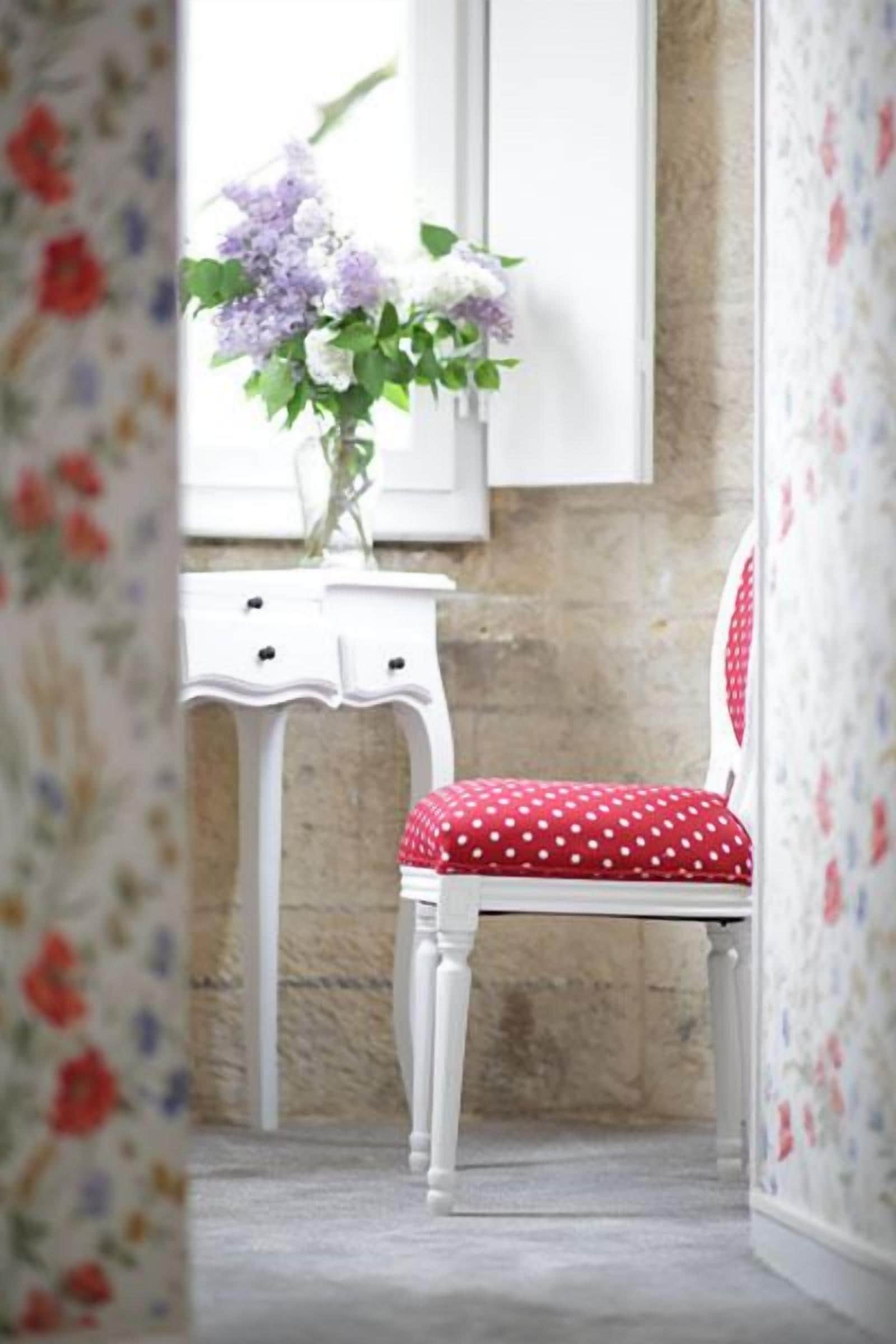 Ein Zimmer mit einem weißen Sessel mit roter Sitzkissen, einer weißen Kommode und einer Blumenvase auf einem Fensterbrett.