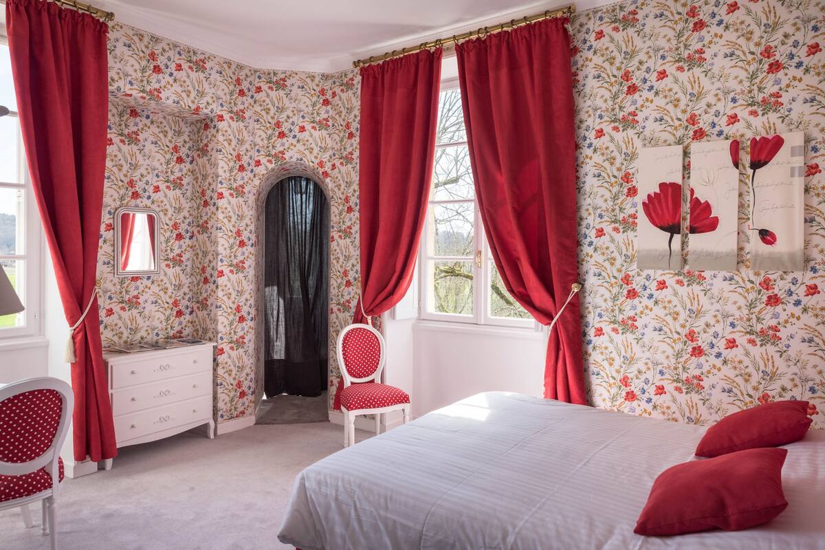 A bedroom with floral wallpaper, a bed with red pillows, a dresser, a chair, and a window with red curtains.