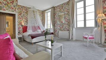 Een hotelkamer met een bed, een zithoek met een stoel, een tafel met een fles en glas, en een groot raam met gordijnen.