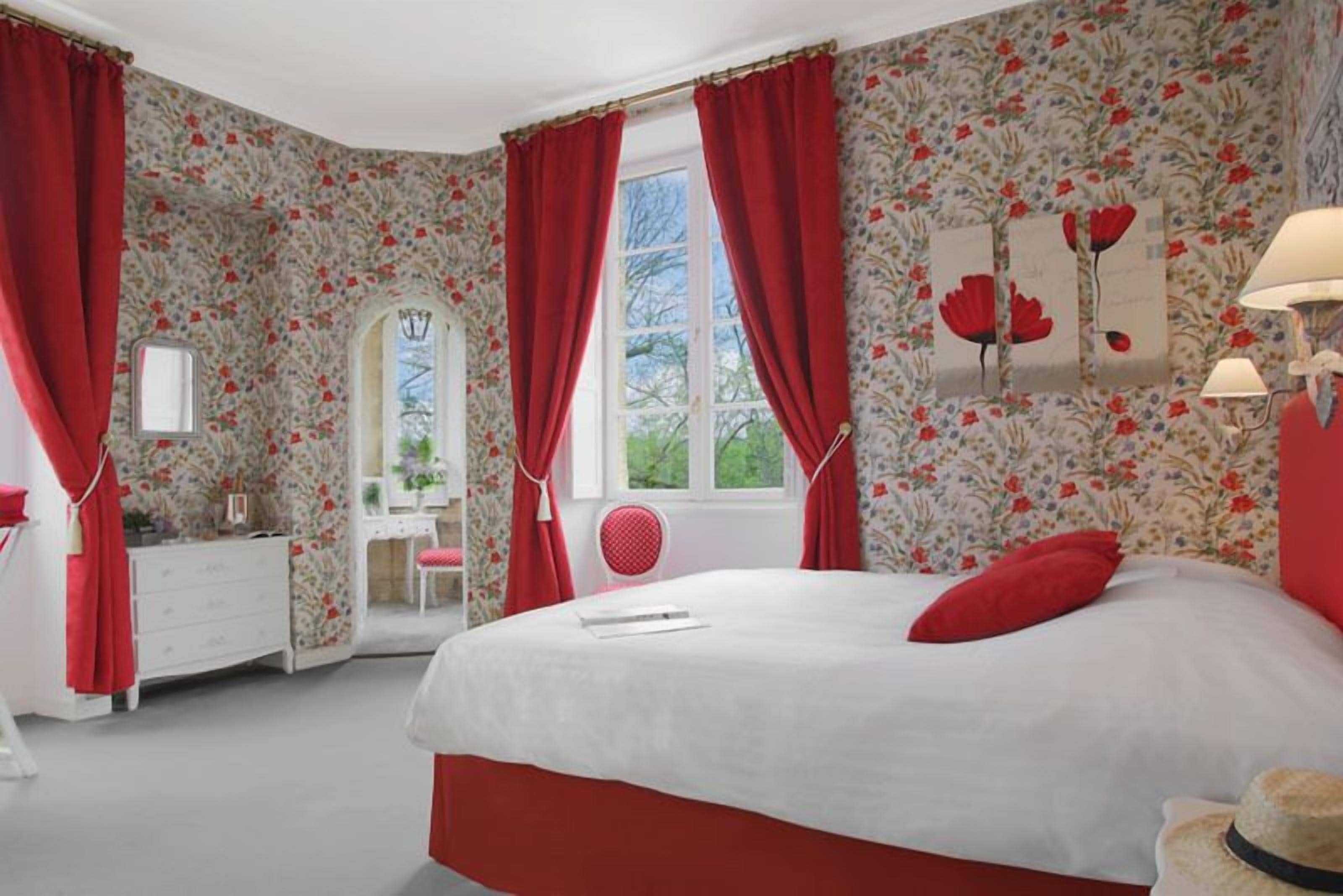 Ein Schlafzimmer mit floralem Wandpapier, einem Bett mit roten Vorhängen, einem Schmuckkästchen, einem Frisiertisch und einem Spiegel.