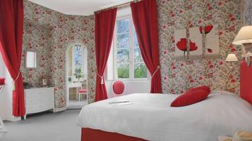 Ein Schlafzimmer mit floralem Wandpapier, einem Bett mit roten Vorhängen, einem Schmuckkästchen, einem Frisiertisch und einem Spiegel.