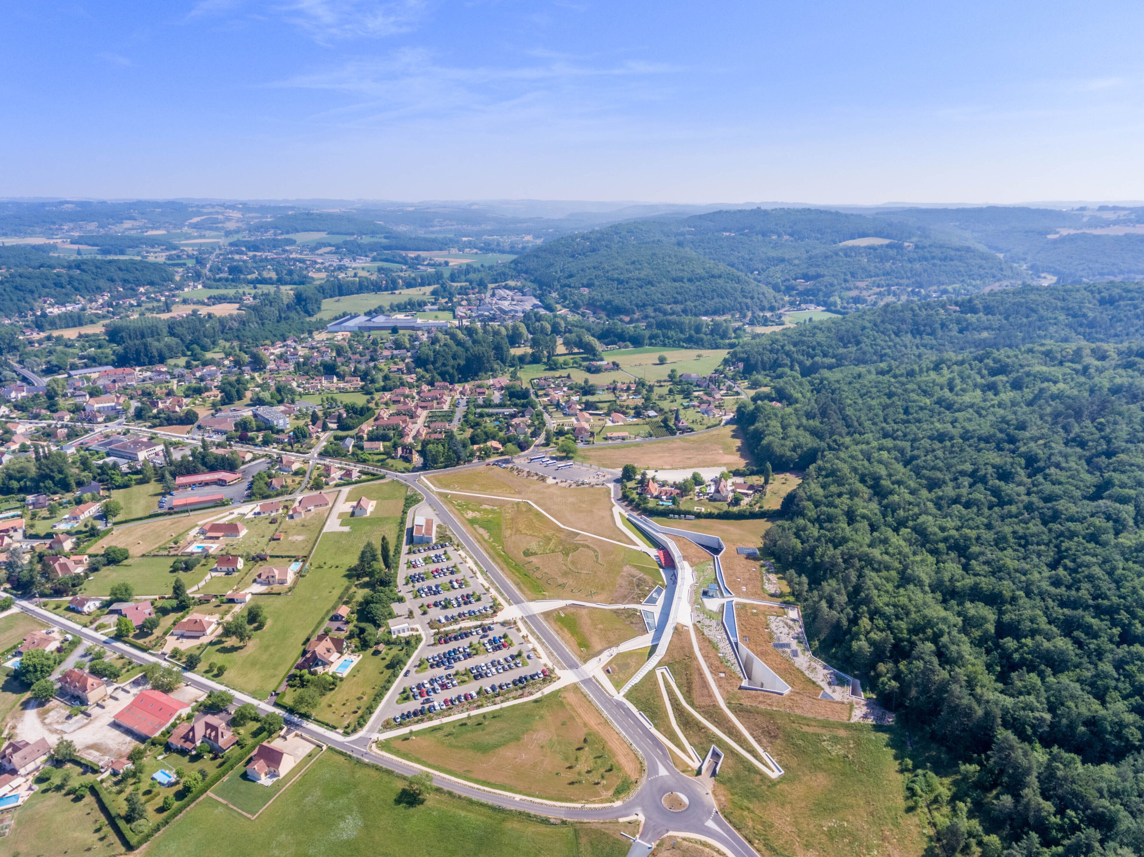 An aerial view of a residential area with a central roundabout, surrounded by houses, green spaces, and a dense forest.