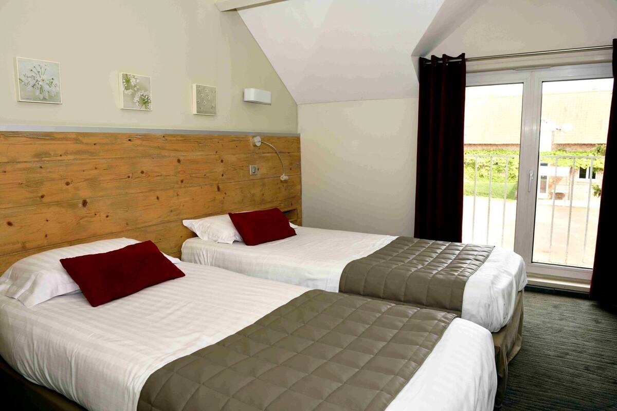 A hotel room with two beds, wooden headboard, white bedding, red pillows, a small wall-mounted light, and a sliding glass door to the outside.