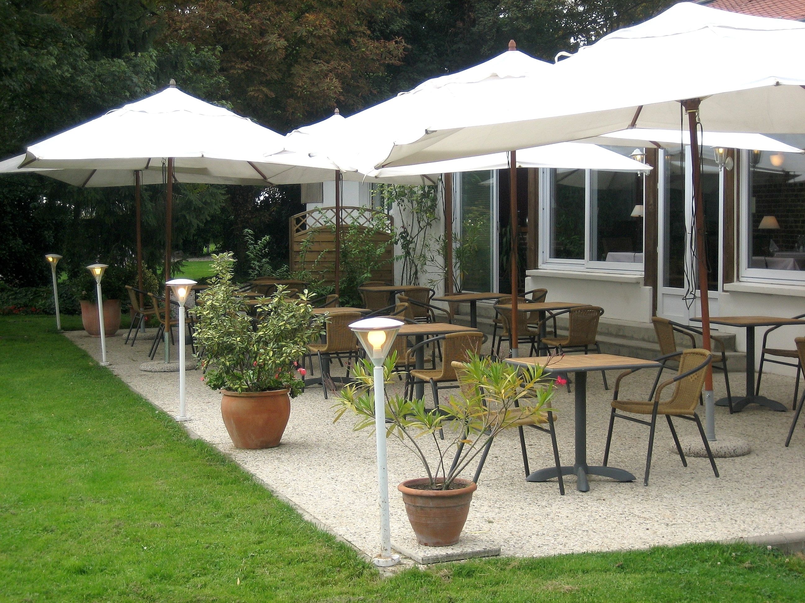 An outdoor patio with multiple tables and chairs under white umbrellas, surrounded by potted plants and outdoor lighting.