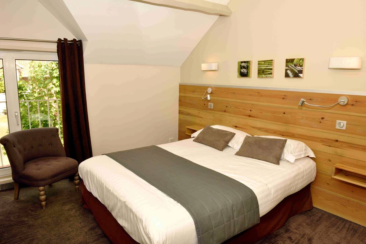 A bedroom with a bed, a chair, wooden headboard, and a window with curtains.