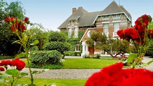 Una casa grande de dos pisos con fachada de ladrillo y techo a dos aguas, rodeada de jardines cuidados y flores rojas en primer plano.
