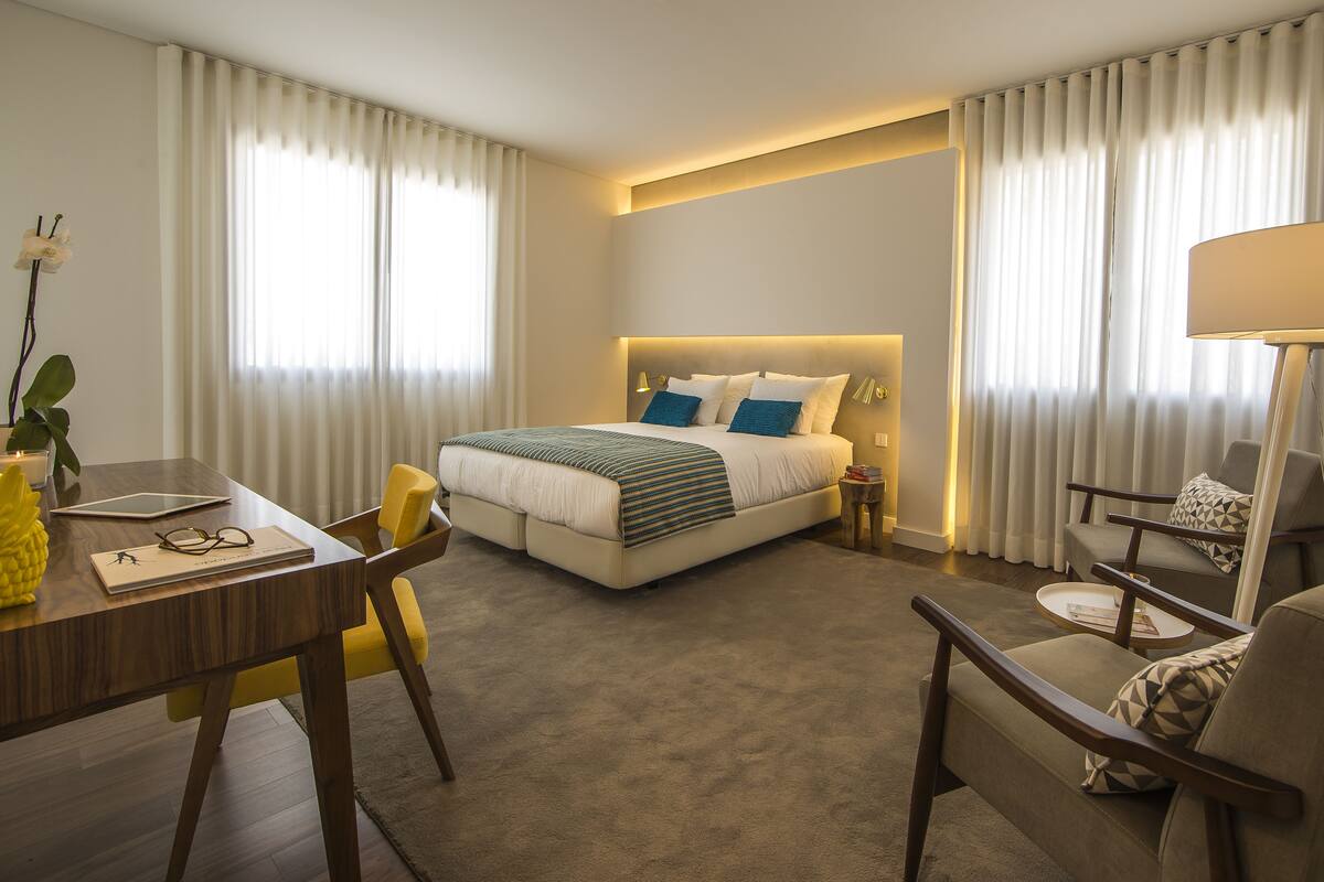 Una habitación de hotel con cama, escritorio, silla, lámpara y ventana con cortinas.