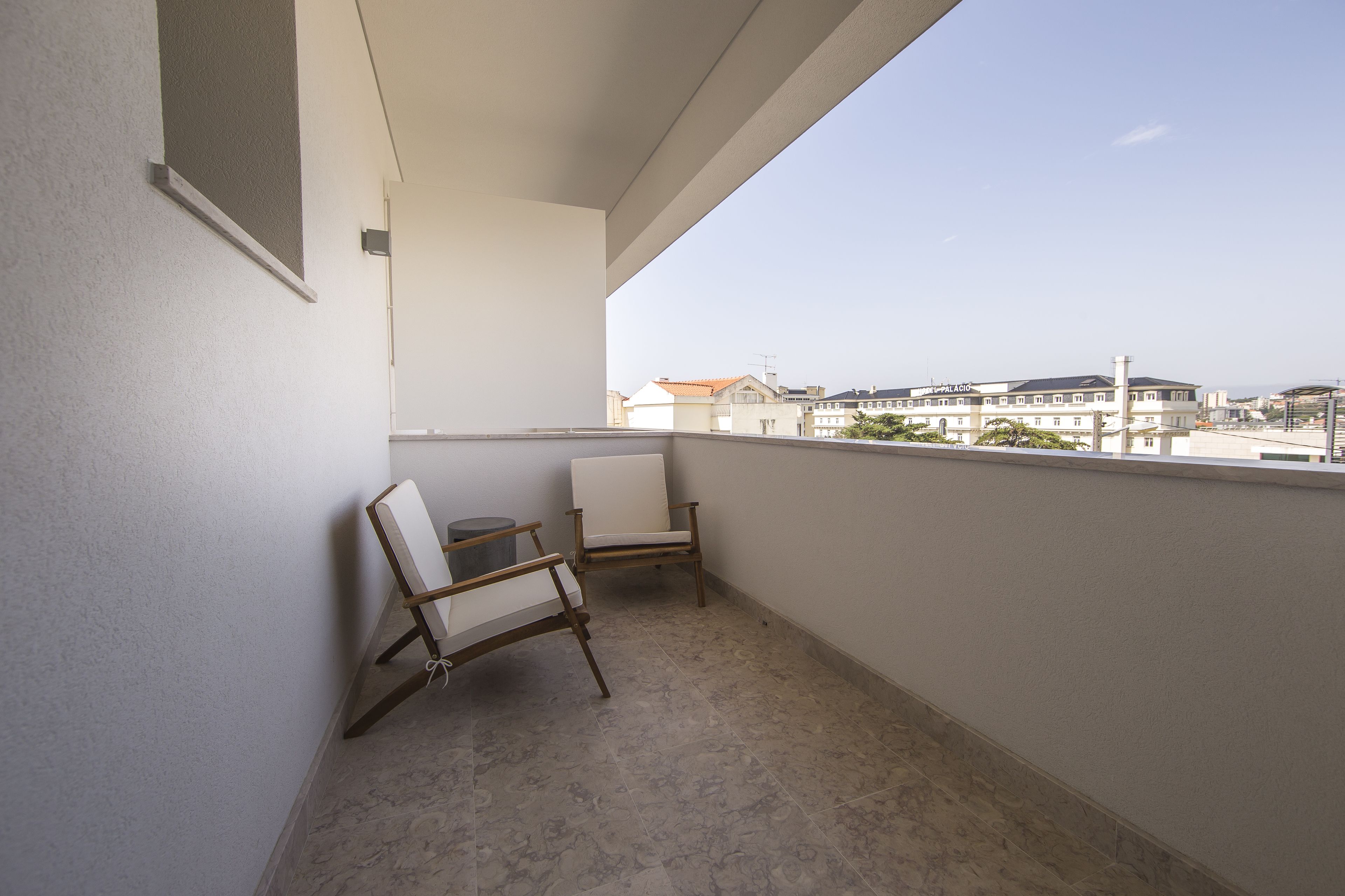 Ein Balkon mit weißem Stuhl und kleinem Tisch, der auf ein Wohngebiet mit Häusern und bewölktem Himmel blickt.
