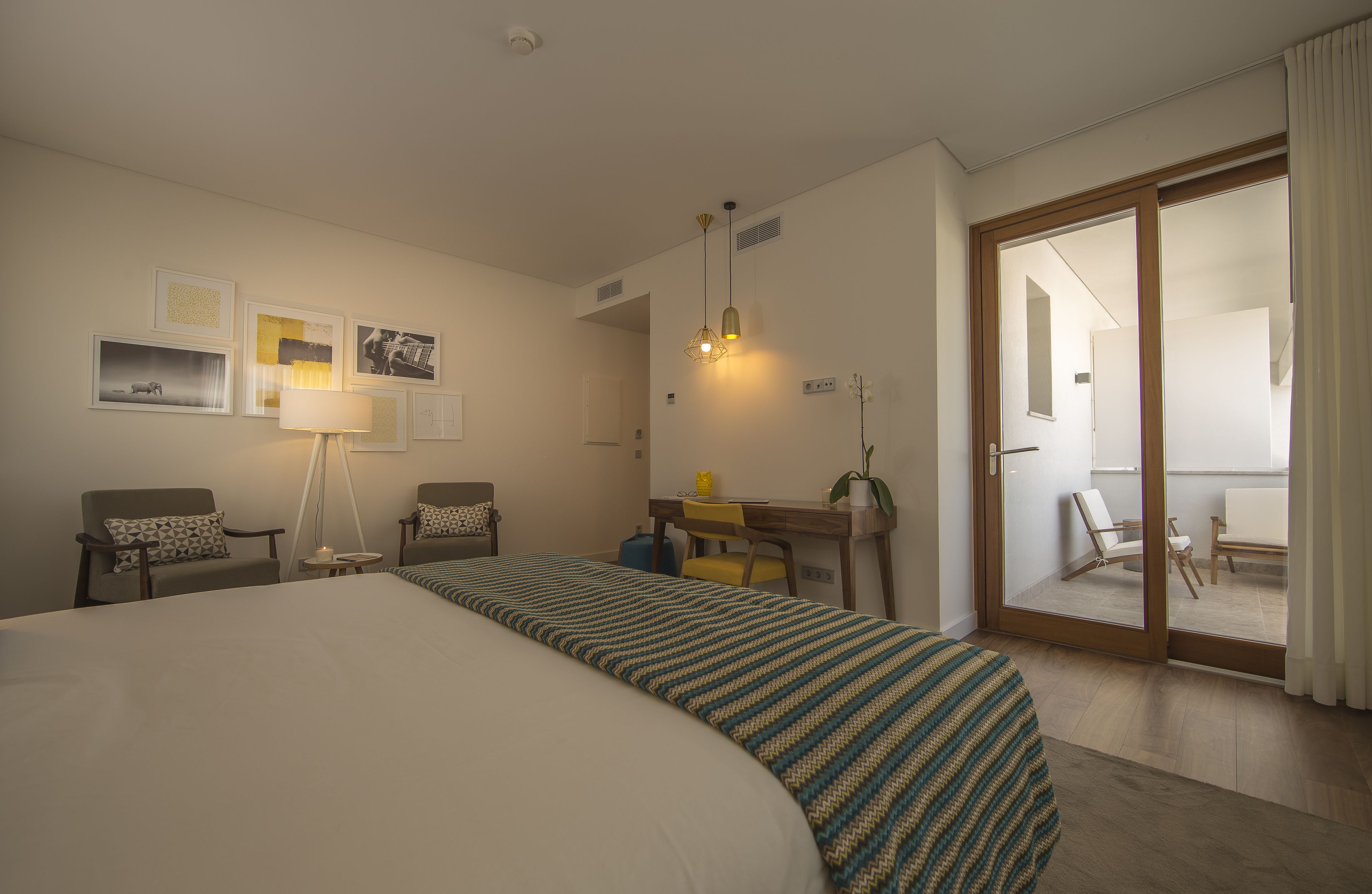 A hotel room with a large bed, two armchairs, a desk with a lamp, and a sliding glass door leading to a balcony.