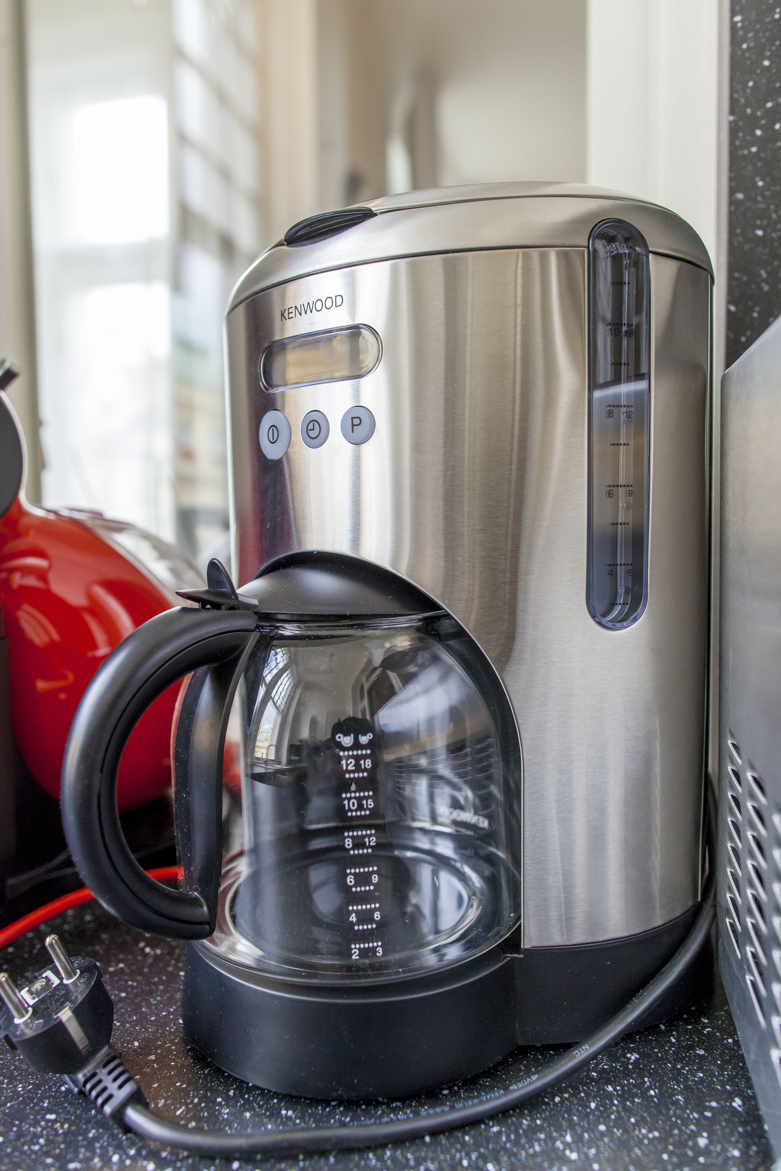 Una cafetera Kenwood con un depósito de agua transparente, panel de control y cable de alimentación sobre una encimera.