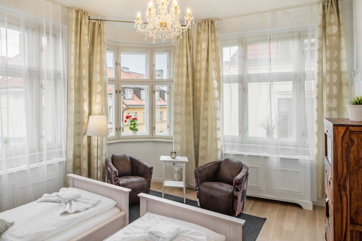 Ein Hotelzimmer mit zwei Betten, einem Chandelier, einem kleinen Tisch mit einer Uhr und Blick auf Gebäude.