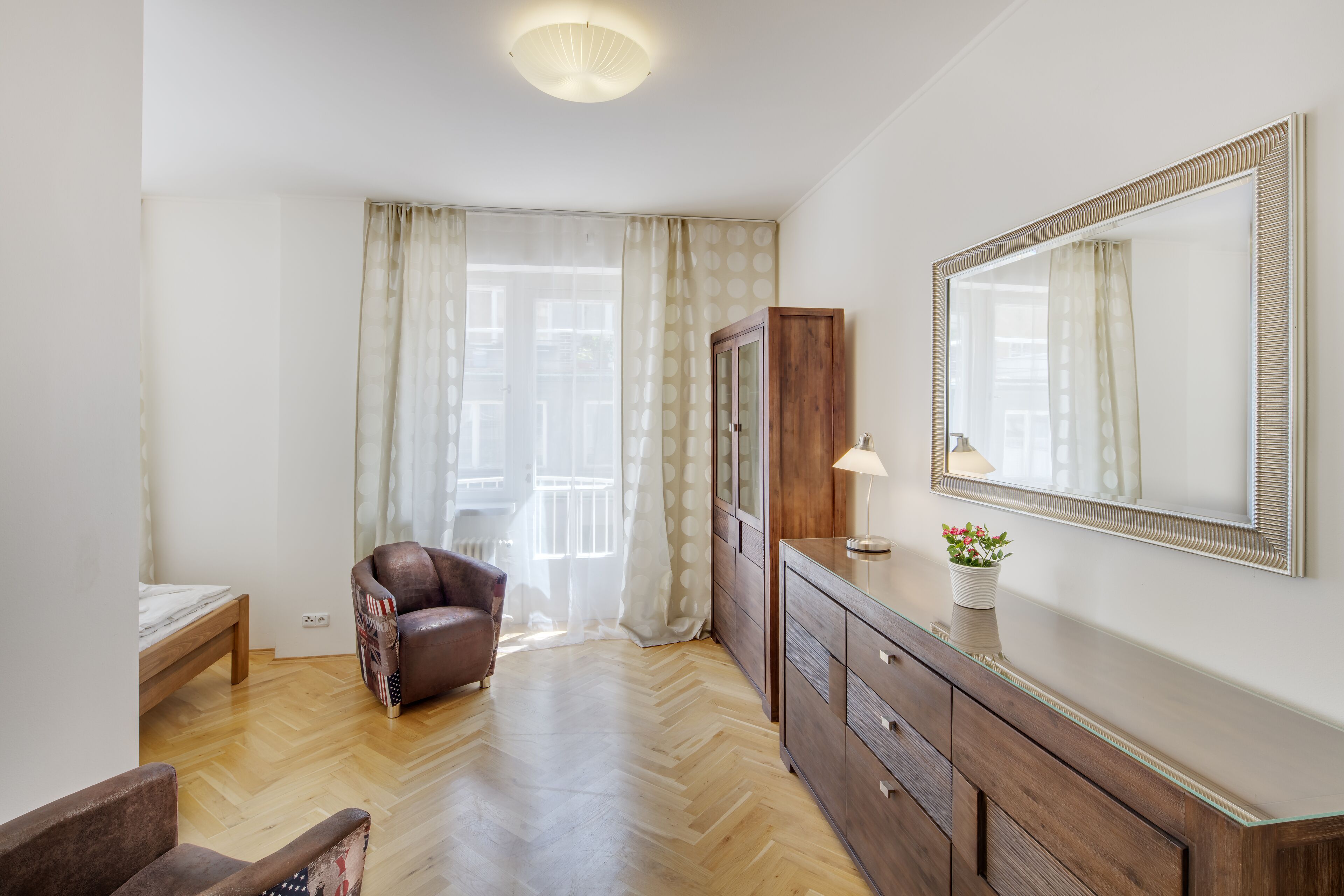 A room with a wooden floor, a brown leather chair, a wooden cabinet, a mirror, and a window with curtains.