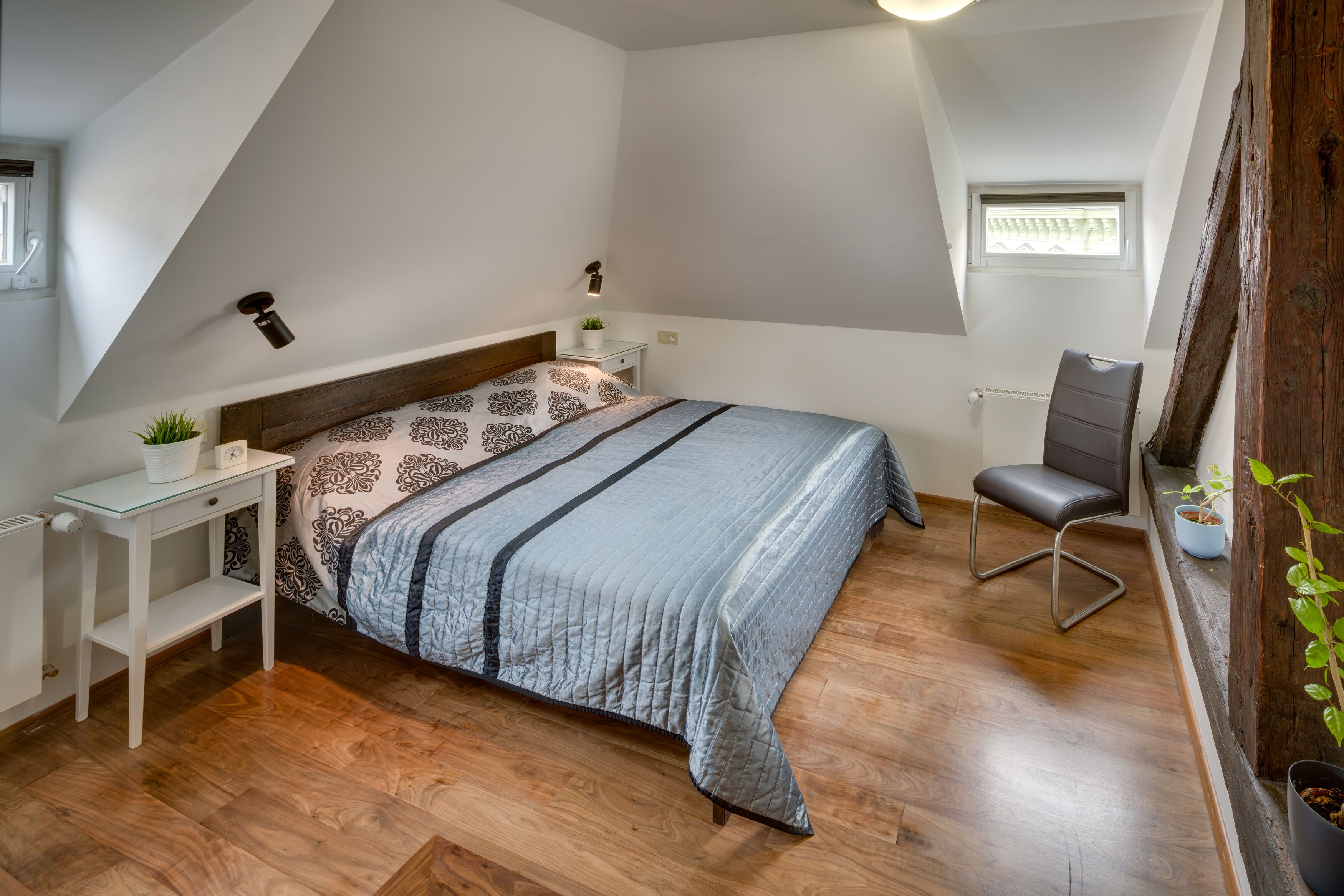 Une chambre avec un plafond en pente, un lit avec une literie à motifs, une table de chevet avec une plante, une chaise et une fenêtre.