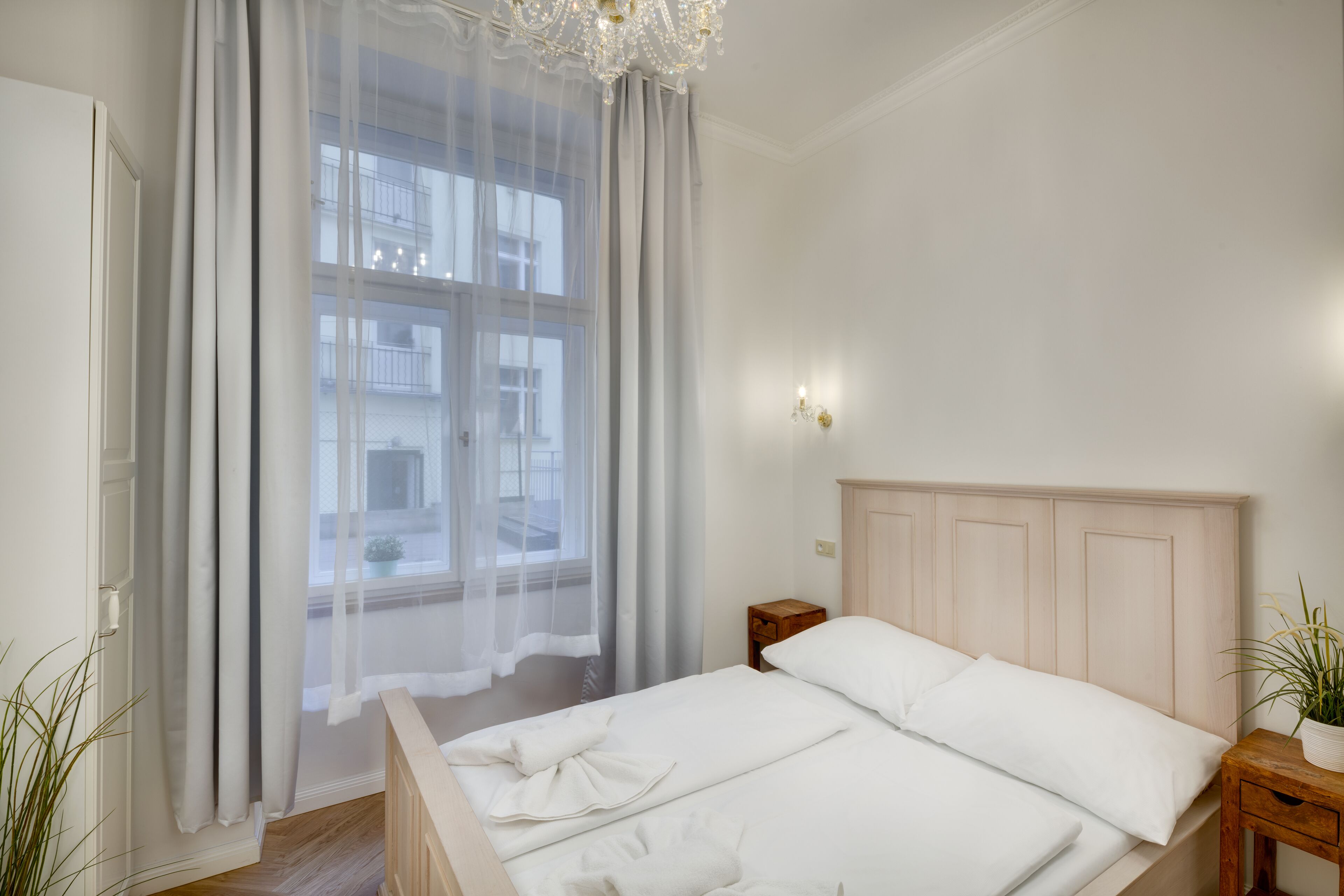 A bedroom with a bed, a nightstand, a window with sheer curtains, and a chandelier.