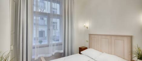 Een slaapkamer met een bed, een nachtkastje, een raam met dunne gordijnen en een kroonluchter.