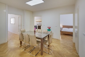 Une chambre d’hôtel moderne avec un plancher en bois, une table à manger en verre et quatre chaises, un lit avec une tête de lit en bois et une petite kitchenette.