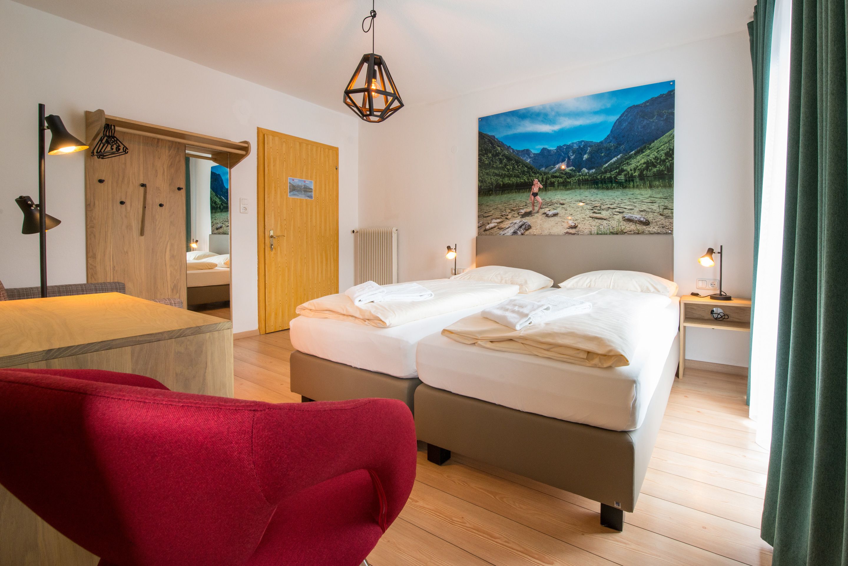 A hotel room with a bed, a red chair, a wooden desk, a wooden door, a wall-mounted lamp, and a large mountain landscape painting.