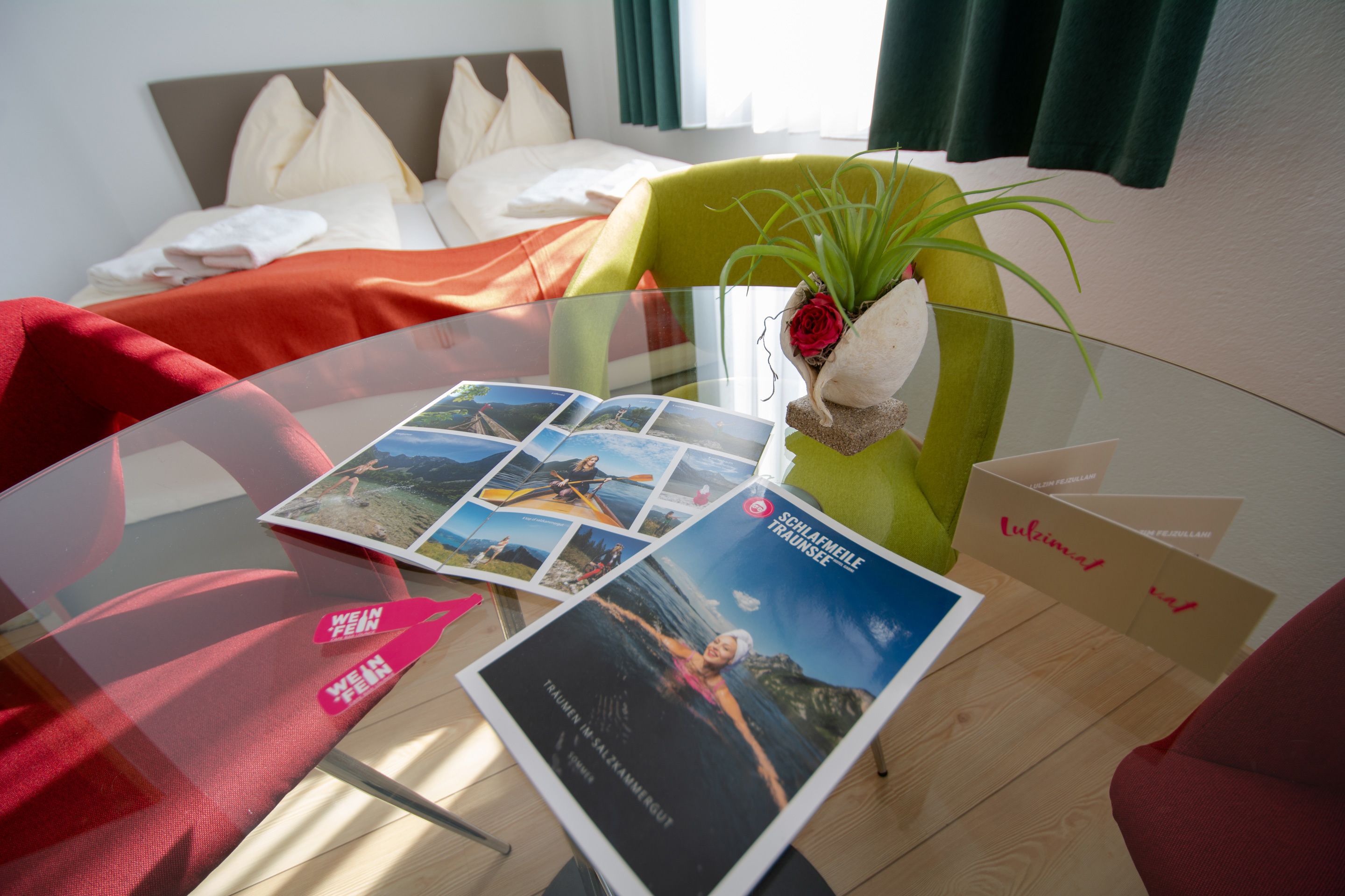 A hotel room with a bed, a glass table with brochures, a green chair, and a potted plant with a rose.