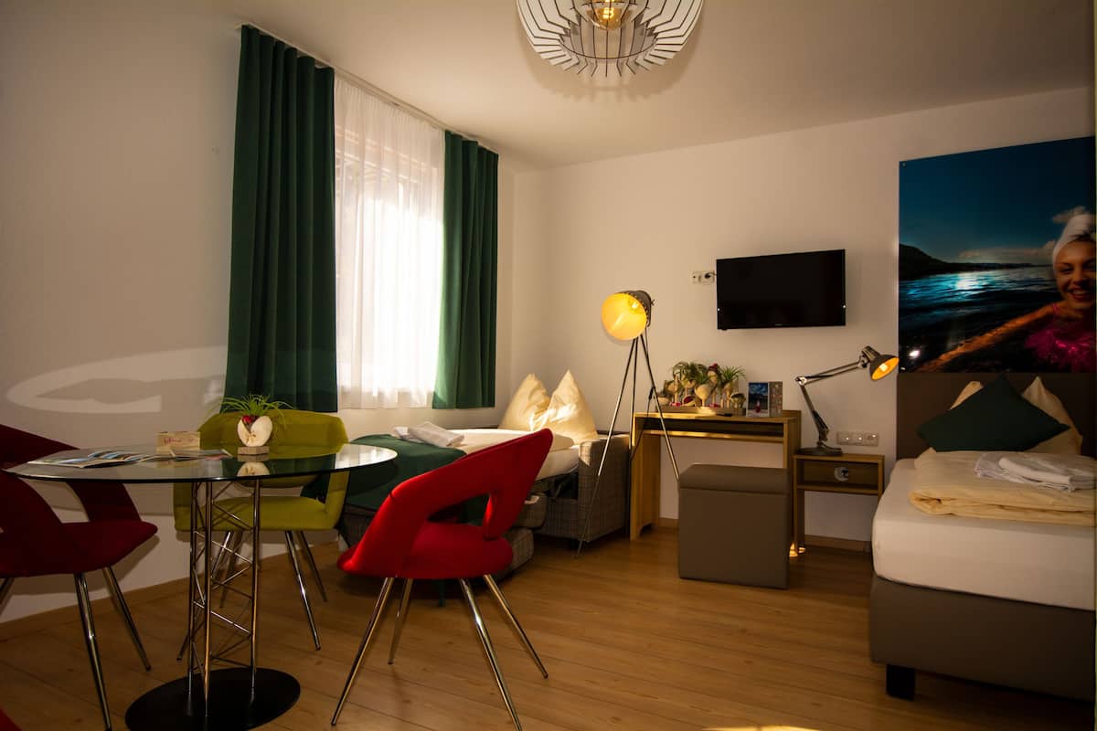 Ein modernes Hotelzimmer mit einem Bett, einem Schreibtisch mit Lampe, einem Fernseher, einem Essbereich mit Stühlen und einem großen Fenster mit Vorhängen.