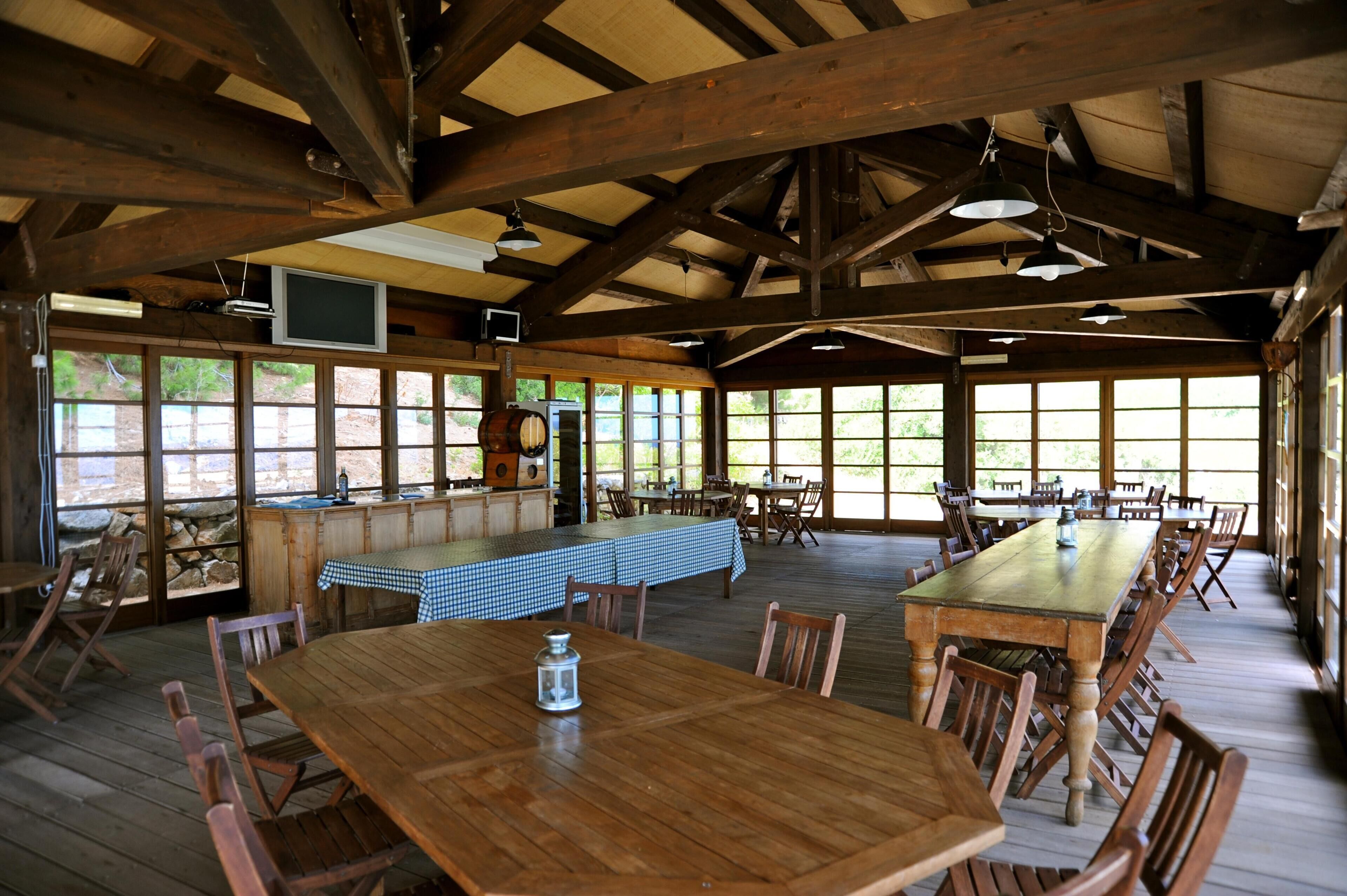 A rustic dining area with wooden tables and chairs, a long table with a checkered tablecloth, and a bar area with a television.