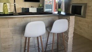 A modern kitchen with a flat-screen TV, two white bar stools, a green vase, and a potted plant.